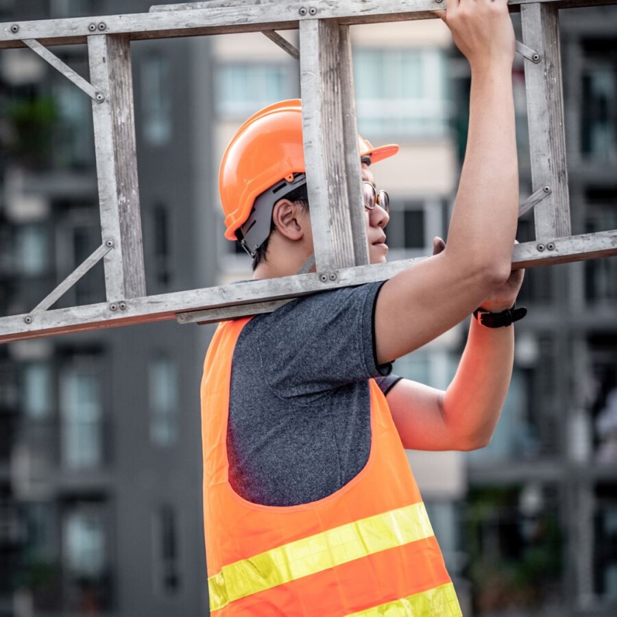 Construction worker wearing a hard hat and high‑visibility vest carrying a ladder on a worksite, representing personal accident and sickness insurance.