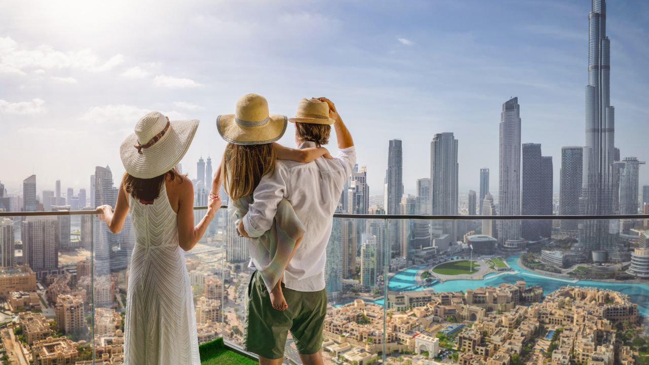 Three people wearing sun hats standing on a balcony overlooking a modern city skyline with tall skyscrapers and a large water feature, representing luxury private client travel insurance.