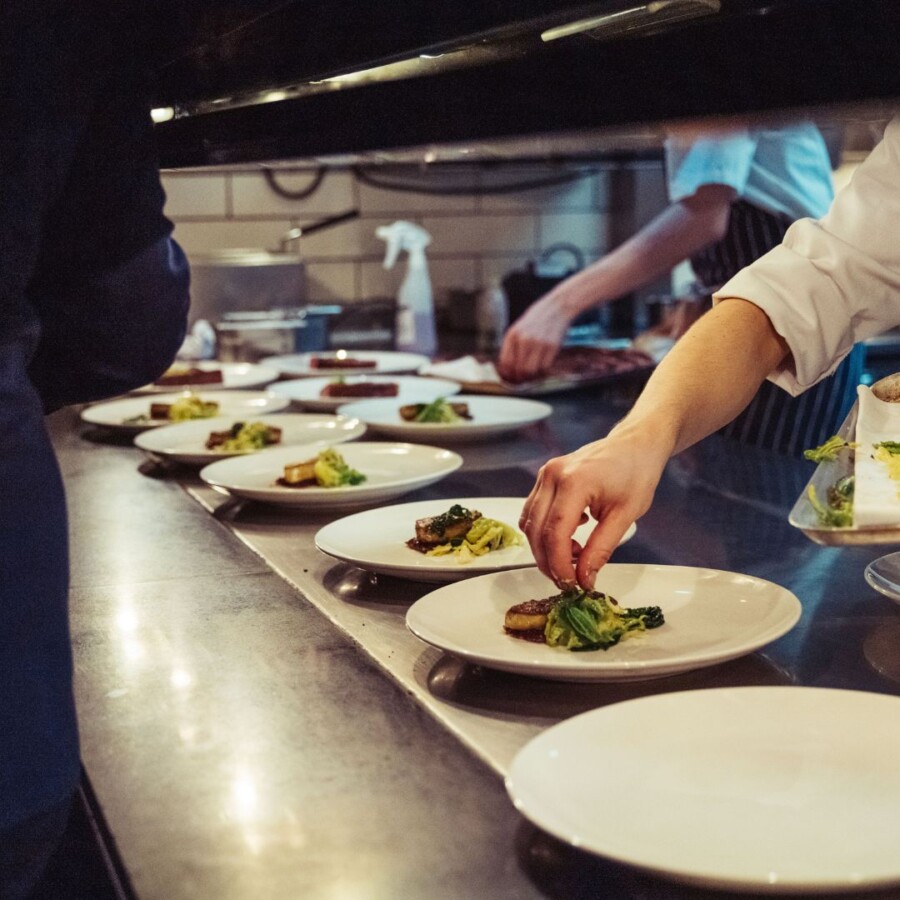 Commercial kitchen pass with multiple plated meals being prepared and arranged by kitchen staff, representing food service environments covered by restaurant, pub and takeaway insurance.
